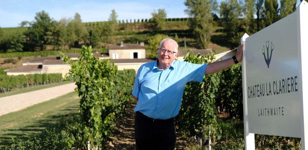 Tony Laithwaite dressed in a short-sleeved blue button down shirt, leans against the welcome sign to Chateau La Clariere. In the background are rows of grape vines.